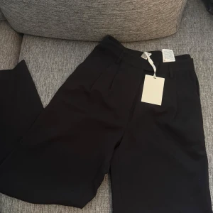 Svarta kostymbyxor från YAS - Snygga svarta kostymbyxor från YAS med klassisk rak passform och hög midja. Byxorna har bälteshällor och pressveck framtill för en clean look. Perfekta att styla med en cool t-shirt eller skjorta. Materialet känns mjukt och har en lätt stretch.