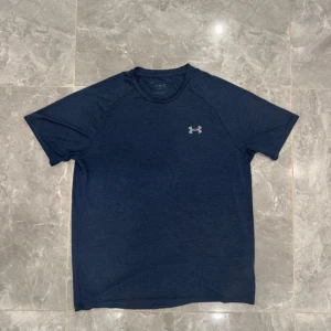 Marinblå t-shirt från Under Armour - Snygg marinblå t-shirt från Under Armour med klassisk logga på bröstet. T-shirten har rund hals och korta ärmar, perfekt för dig som gillar sportig stil. Materialet är mjukt och andas, vilket gör den grym för träning eller chill.