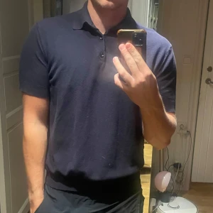 Mörkblå silk blend pikétröja These Glory Days - Säljer en stilren mörkblå pikétröja från These Glory Days i silk blend-material. Tröjan har klassisk krage, korta ärmar och svinläckra knappar framtill. Perfekt för dig som gillar en clean look med lite lyxig känsla. Passar dig som vill ha något bekvämt men ändå snyggt. Oanvänd 