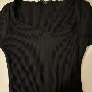 Svart ribbad topp - Svart ribbad topp i storlek XS. Toppen har en snygg v-ringad hals och korta ärmar. Materialet är mjukt och stretchigt, vilket gör den bekväm att bära. Perfekt för dig som gillar stilrena och enkla plagg med lite edge.