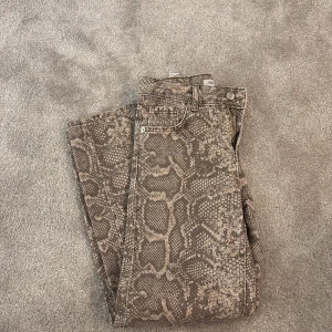 Ormmönstrade jeans från Zara - Säljer ett par sjukt coola jeans från Zara med ormmönster i beige och bruna toner. Klassisk femficksmodell med knapp och dragkedja framtill. Materialet är jeans och passformen är normal, perfekt för dig som vill sticka ut med din stil.