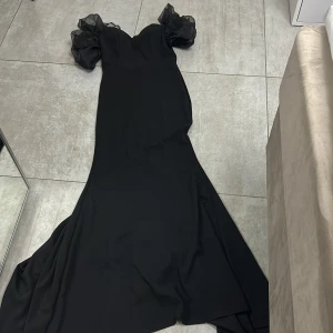 Svart långklänning med puffärmar - Säljer en elegant svart långklänning med dramatiska puffärmar i transparent tyg. Klänningen har en figurnära siluett upptill och en svepande kjol som ger en riktigt snygg look. Perfekt för dig som vill sticka ut med stilrena detaljer och volym vid axlarna.