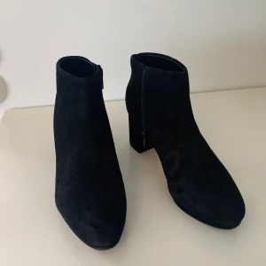 Svarta mocka stövletter från Clarks stl 37,5 - Snygga svarta stövletter från Clarks i mjuk mocka med rund tå och stabil blockklack. Skorna har dragkedja på sidan för enkel på- och avtagning. Perfekt till höstens och vinterns outfits när du vill ha en stilren look. Endast provade utomhus vid ett tillfälle så i absolut nyskick. OBS storlek 37,5