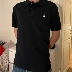 Svart Polo Ralph Lauren t-shirt - Svart t-shirt från Polo Ralph Lauren med klassisk pikékrage och två knappar framtill. Slim fit-modell med korta ärmar och ikonisk vit logga broderad på bröstet. Tillverkad i mjuk bomull som är skön mot huden. Perfekt för en clean och stilren look. Passar  storlek large tycker inte de är custom slim tycker den är som en vanlig Large är billigare vid snabbaffär den är inte andvänd så mkt endast i få till fällen