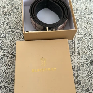 Burberry rutig bälte med guldfärgat spänne - Snyggt Burberry-bälte med klassiskt rutigt mönster i beige, svart och röd. Bältet har ett exklusivt guldfärgat spänne med Burberrys ikoniska ryttare. Materialet är skinn med textilstruktur och bältet levereras i original ask.