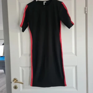 Svart klänning med röda ränder NLY Trend - Svart kortklänning från NLY Trend i storlek M. Klänningen har röda och vita ränder längs sidorna och på ärmarna, vilket ger en sportig vibe. Modellen är figurnära med rund hals och korta ärmar. Materialet känns mjukt och stretchigt, troligen bomull eller polyester.