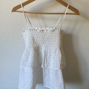 Vit blus med volanger från Gina Tricot - Supersöt vit blus från Gina Tricot med tunna axelband och smock upptill. Blusen har volanger och små guldiga prickar som ger extra glans. Perfekt för sommarens alla häng! Lätt och luftig i bomullsmaterial.