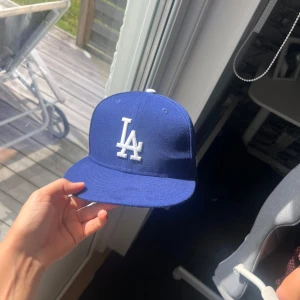 Blå LA Dodgers keps från New Era - Snygg blå keps från New Era med LA Dodgers-logga broderad i vitt framtill. Klassisk modell med rak skärm och broderade ventilationshål. MLB-logga och New Era-märke på sidan. Perfekt för dig som vill ha sportig streetstyle. 