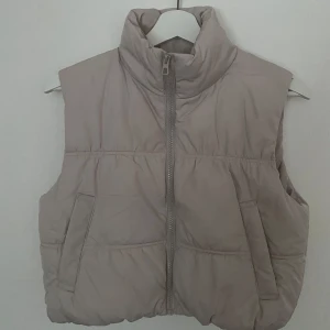 Beige dunväst från H&M Divided XS - Säljer en beige kroppar dunväst från H&M Divided i storlek XS. Västen har hög krage, dragkedja framtill och två snedställda fickor. Den är quiltad och har en clean, enkel look som passar till många outfits.