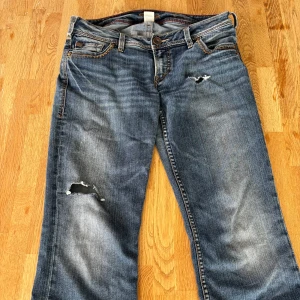 Blå bootcut jeans med slitningar - Säljer ett par blå bootcut jeans med låg midja och slitna detaljer på benen. Jeansen har klassisk femficksdesign, dragkedja och knapp framtill. Materialet är jeans med snygg tvätt och slitningar för en cool, avslappnad vibe. Innerbenslängd 79 & midjemått 38 💙
