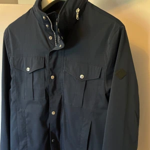 Marinblå fieldjacket från J Lindeberg  - Snygg marinblå fieldjacket från J Lindeberg med hög krage, silverfärgade knappar och dragkedja. Jackan har två stora bröstfickor med lock och emblem på ärmen. Perfekt för dig som gillar stilren och funktionell design. Skick 8/10 