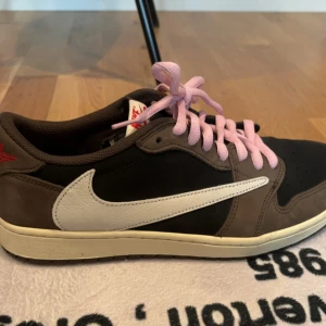 Nike Jordan 1 Low Travis Scott - Säljer ett par Nike Jordan 1 Low Travis Scott med bruna och svarta paneler i mocka, vit omvänd swoosh på utsidan och klassisk swoosh på insidan. Skorna har både svarta och ljusrosa snören, samt detaljer med röd text. Perfekt för dig som vill sticka ut med hypade sneakers. I storlek 11 (EU 45). Aldrig använda