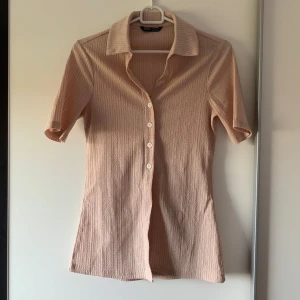 Beige topp  - Snygg beige topp med ribbad struktur och korta ärmar. Toppen har klassisk krage, vita knappar framtill och är figurnära i modellen. Materialet är mjukt och stretchigt, perfekt för en chill och stilren look.