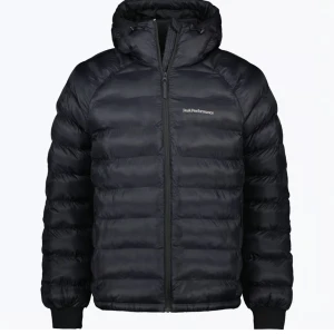 Svart dunjacka Peak Performance - Snygg svart dunjacka från Peak Performance med huva och diskret logga på bröstet. Jackan har quiltad design, dragkedja framtill och ribbade muddar vid ärmslut. Perfekt för kalla dagar och har en clean, sportig look som funkar till streetwear.