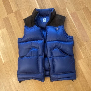 Blå dunväst Wrangler Blue Bell M - Säljer en blå dunväst från Wrangler Blue Bell i storlek M. Västen har bruna axelpartier i mocka, hög krage och två stora snedställda fickor framtill. Stängs med dragkedja och har en klassisk, puffig look som är riktigt snygg till hoodie eller skjorta.