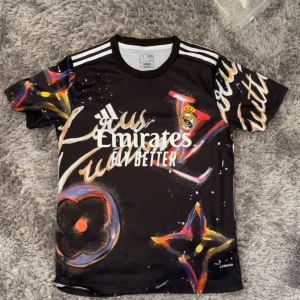 Real Madrid svart fotbollströja - Svart fotbollströja från Adidas med Real Madrid-logga och Emirates-tryck på bröstet. Tröjan har coolt grafiskt mönster i färger som röd, blå, gul och orange. Klassiska vita ränder på axlarna och rund hals. Materialet är lätt och andas.
