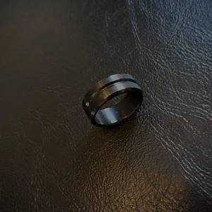 Svart bred ring med cool design - Size 10.0-10.5 Säljer en svart ring med bred form och en snygg upphöjd detalj runt hela ringen. Den har en modern och stilren look som passar perfekt om du gillar minimalistiska accessoarer. Ringen är i metall och känns riktigt robust på fingret.