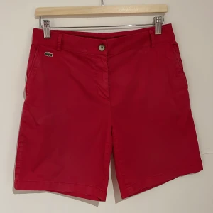 Röda shorts från Lacoste, strl 38 - Snygga röda shorts från Lacoste med klassisk krokodillogga vid fickan. Modellen har midwaist, raka ben och bälteshällor. Materialet är mjuk bomull. Perfekta för sommaren och har stilrena fickor både fram och bak.