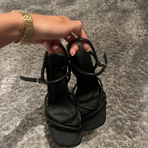 Svarta sandaler med klack från Skopunkten - Säljer ett par svarta sandaler från Skopunkyen med smala remmar och fyrkantig tå. Skorna har en elegant låg klack och justerbart spänne vid ankeln. Materialet är skinn och designen är stilren med en modern vibe, perfekt för dig som gillar minimalistiska detaljer.