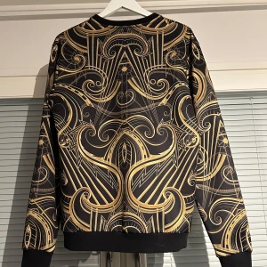 Versace Jeans långärmad tröja – svart med guldmönster, storlek L - Snygg svart tröja från Versace Jeans, med fett guldmönster, Pris kan diskuteras vid snabb affär. Hybrid mellan M/L Passar både perfekt, går inte att köpa denna vara längre i butik då det är en limited edition vara.