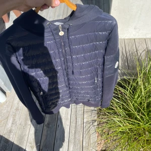 Mörkblå Moncler cardigan  - Säljer en mörkblå pufferjacka från Moncler med huva och snygga quiltade detaljer på framsidan. Jackan har dragkedja, två fickor med zip och Moncler-logga på ärmen. Materialmix med polyester och bomull, perfekt för dig som gillar streetwear. Har kvitto och tags och allt 