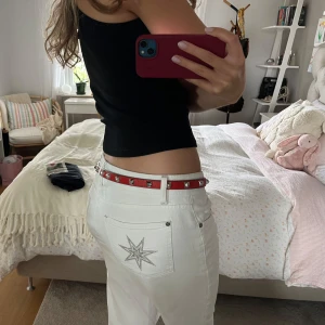 Rött skärp med nitar från Karen Millen - Snyggt rött bälte från Karen Millen med silverfärgade fyrkantiga nitar längs hela remmen. Bältet är smalt och har en klassisk metallspänne. Tillverkat i skinn med svarta detaljer på insidan. Perfekt för att ge din outfit en edgy touch.