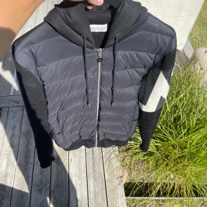 Svart Moncler cardigan  - SÄLLSYNT Snygg svart pufferjacka från Moncler med stickade ärmar och huva. Jackan har dragkedja framtill, snörning vid huvan och ett coolt vitt zigzag-mönster på ärmarna. Perfekt för dig som gillar streetstyle och exklusiva märken.