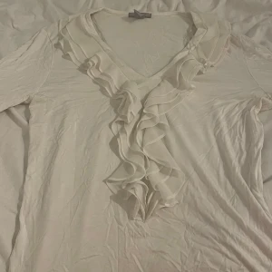 Vit blus med volang från H&M - Supersöt vit blus från H&M i storlek L med v-ringning och snygga volanger längs halsen och framtill. Långärmad och tillverkad i mjuk bomull, perfekt för dig som gillar romantiska detaljer och vill ha en chill men ändå stylish look.