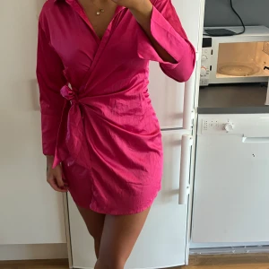 Kort rosa omlottklänning med knyt - Säljer en snygg kort rosa klänning med omlott-design och knyt i sidan. Klänningen har långärmade ärmar med breda manschetter och en krage som ger en cool skjortkänsla. Materialet är glansigt och känns lätt mot huden, perfekt för dig som gillar att sticka ut.