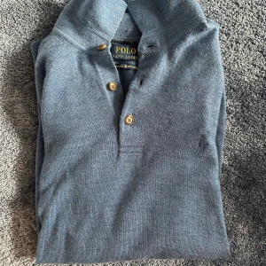 Blå merinoull polotröja Polo Ralph Lauren - Snygg blå polotröja från Polo Ralph Lauren i tvättbar merinoull. Tröjan har klassisk krage, tre knappar framtill och broderad logga på bröstet. Långärmad och perfekt för dig som gillar stilren och tidlös design. 