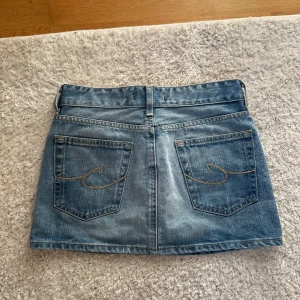 Blå jeanskjol med bakfickor - Snygg blå jeanskjol med klassisk femficksdesign och dubbla knappar framtill. Kjolen har två stora bakfickor med dekorativ söm och bälteshällor i midjan. Perfekt för en avslappnad och trendig look.