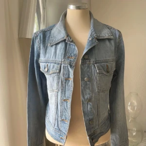 Ljusblå jeansjacka med knappar - Snygg ljusblå jeansjacka med klassisk krage och silverfärgade knappar framtill. Jackan har två bröstfickor med lock och knapp, samt justerbara detaljer vid midjan. Tillverkad i 100% bomull för en skön och avslappnad känsla. Perfekt för dig som gillar en tidlös streetstil.