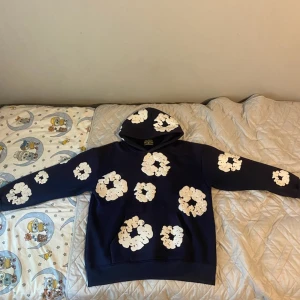 Blå hoodie med vita blommor Denim Tears - Säljer en blå hoodie från Denim Tears med stora vita blommönster över hela tröjan, även på huvan. Tröjan har klassisk känguruficka och huva. Perfekt för dig som vill sticka ut med en unik och kreativ stil.