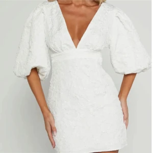 Vit kort klänning med puffärmar - Supersnygg vit kort klänning med djup v-ringning och stora puffärmar. Klänningen har en markerad midja och är gjord i ett strukturerat tyg som ger en lyxig känsla. Perfekt för dig som vill sticka ut med en trendig och feminin look.