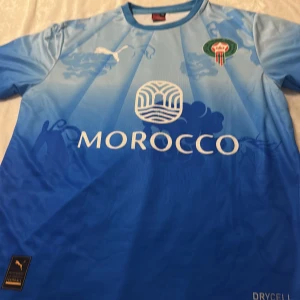 Blå Marocko fotbollströja Puma - Snygg blå Marocko fotbollströja från Puma med FRMF-emblem på bröstet och vit Puma-logga. Tröjan har korta ärmar, rund hals och coola grafiska detaljer på framsidan. Materialet är lätt och andas, perfekt för match eller träning. DryCell-teknologi för extra komfort.