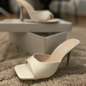 Vita klackar  - Snygga vita klackar med öppen tå och hög klack. Skorna har en fyrkantig tå och ett stilrent croco-mönster på ovandelen. Perfekt för dig som gillar minimalistisk design med en twist. Materialet känns som syntet och ger en lyxig känsla. Helt nya. Oanvända. 