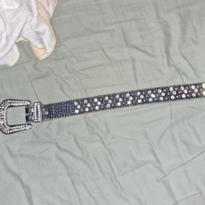 Svart bb simon bälte med nitar och strassdetaljer - Säljer ett svart bb simon bälte med glittriga strassdetaljer och silvriga nitar. Bältet har ett stort, dekorerat spänne och är perfekt för att ge outfiten lite extra edge. Märkt med 'Fashion Belt' på metallplatta.