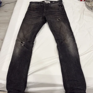 Svarta Replay jeans med slitningar - Säljer ett par svarta Replay jeans med snygga slitningar och diskret tvättad look. Byxorna har klassisk femficksdesign, dragkedja och knapp i midjan samt raka ben. Perfekta för dig som gillar en lite tuffare stil.