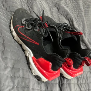 Nike React svarta och röda sneakers - Snygga Nike React sneakers i svart med röda detaljer och vit sula. Skorna har ventilerande mesh-ovandel, röda accenter på hälen och snörning framtill. Perfekta för dig som gillar sportig och modern stil.