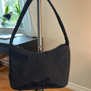 Svart axelväska i syntetmaterial - Enkel och stilren svart axelväska med mjukt, vadderat handtag och dragkedja upptill. Väskan har en minimalistisk design och är tillverkad i slätt syntetmaterial. Perfekt storlek för det viktigaste och passar till många olika outfits.