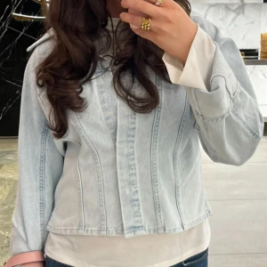 Ljusblå croppad jeansjacka - Snygg ljusblå jeansjacka med croppad passform och markerade sömmar. Jackan har klassisk krage, långa ärmar och knäppning framtill. Perfekt för dig som vill ha en stilren och trendig look till vardags.