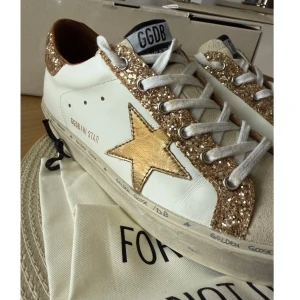 Golden Goose sneakers med glitter och stjärna - Snygga Golden Goose sneakers i vitt skinn med guldglittriga detaljer och en metallic guldstjärna på sidan. Skorna har vit snörning, beige mockadetaljer och platt sula. Perfekta för dig som vill sticka ut med en cool och lyxig look. Köpt på vestiaire 