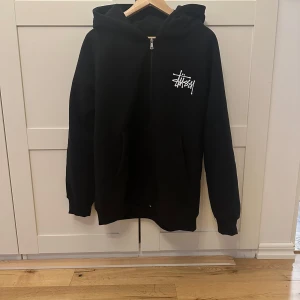 Svart hoodie från Stüssy - Säljer en svart hoodie från Stüssy i storlek M. Tröjan har dragkedja, huva och fickor framtill samt Stüssy-logga tryckt på både bröstet och ryggen. Perfekt för en avslappnad streetwear-look.