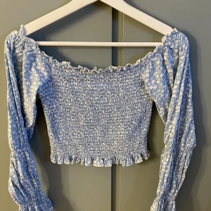 Ljusblå croppad blus med blommigt mönster från NA-KD - Supersöt ljusblå croppad blus från NA-KD med små vita blommor, smockad överdel och volangdetaljer vid halsringning och nederkant. Långa puffärmar med resår vid handlederna ger en romantisk känsla.