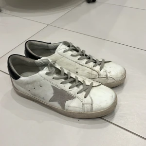 Vita sneakers med grå stjärna från Golden Goose - Säljer ett par vita sneakers från Golden Goose med grå stjärna på sidan och svart häl. Skorna har snörning och platt sula, samt klassisk rund tå. Perfekta för dig som gillar en stilren och trendig look. Dom har blivit för små för mig och vill bli av med dom pris kan diskuteras