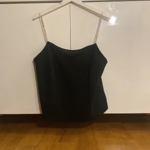 Svart linnetopp med glittriga axelband - Snygg svart linnetopp med tunna glittriga axelband. Toppen har en enkel och stilren design med rak halsringning och något lös passform. Perfekt att styla till olika outfits för en trendig look.