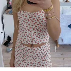 Vit blommig kortkjol med volang - Supersöt vit kortkjol med små röda blommor och gröna blad. Kjolen har en volangkant längst ner som ger en gullig och somrig vibe. Perfekt för dig som gillar romantiska och lekfulla plagg. Materialet känns lätt och luftigt.