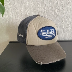 Trucker keps från Von Dutch - Snygg keps från Von Dutch i beige och brunt med broderad logga framtill. Kepsen har en justerbar rem baktill och en sliten look med en liten reva på skärmen. Perfekt för en avslappnad stil.                   Pris kan diskuteras!