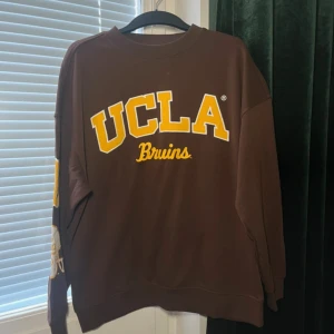 Brun UCLA sweatshirt från H&M Divided - Brun sweatshirt från H&M Divided med stor gul UCLA Bruins-tryck på bröstet och coolt tryck med hjälm och siffror på ena ärmen. Oversized fit och rund halsringning, perfekt för en avslappnad college-look.