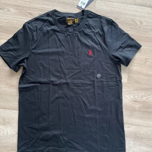 Svart t-shirt Polo Ralph Lauren M - Svart t-shirt från Polo Ralph Lauren i bomull med klassisk rund hals och röd broderad logga på bröstet. Modellen är slim fit och har korta ärmar. Tröjan är helt oanvänd endast testad. Den har legat i min garderob den senaste månaden men skicket är fortfarande samma som när den köptes! Pris kan diskuteras vid en snabb affär